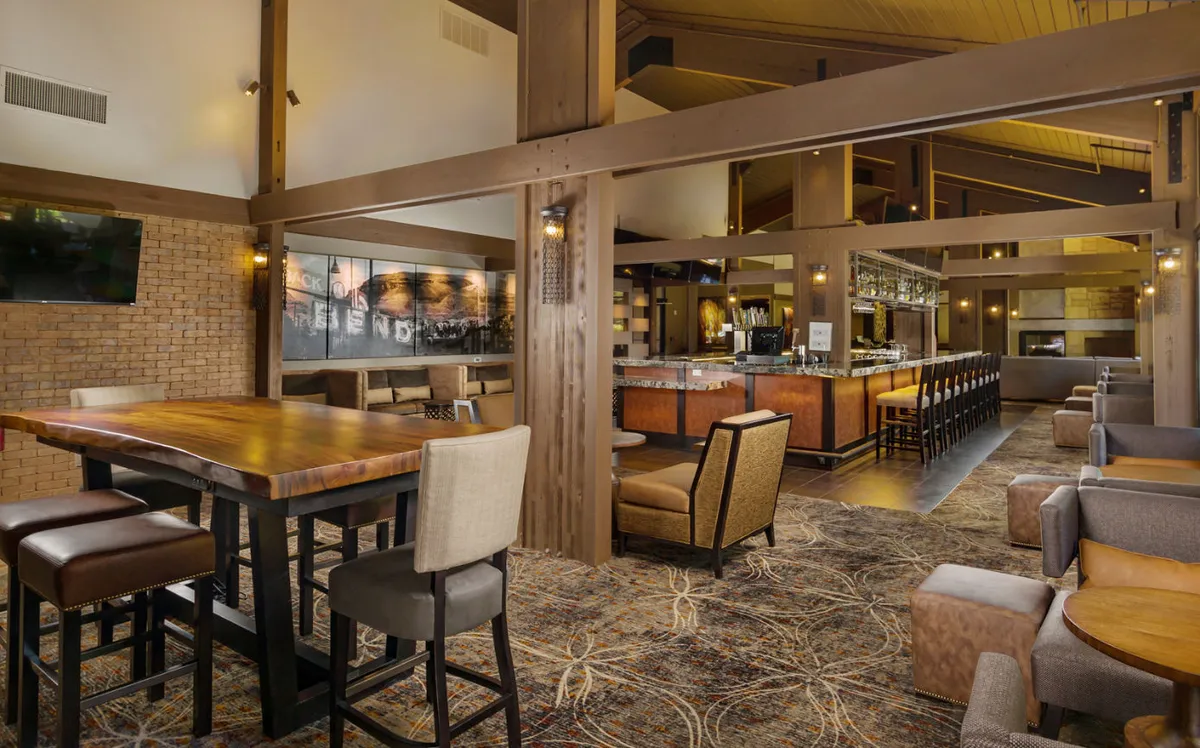 A spacious interior view featuring a large wooden communal table, a well-lit bar area with an array of bottles, and comfortable seating options. Guests are seen socializing, dining, and relaxing with drinks, contributing to a lively yet inviting atmosphere. The setting is ideal for gatherings, group chats, or unwinding after a long day.