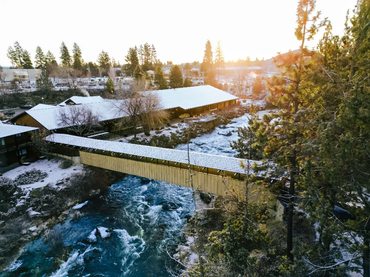 A covered bridge spans the flowing Deschutes River, bordered by trees and snow-dusted rooftops. The serene atmosphere and inviting path suggest effortless access to nature and year-round adventure.