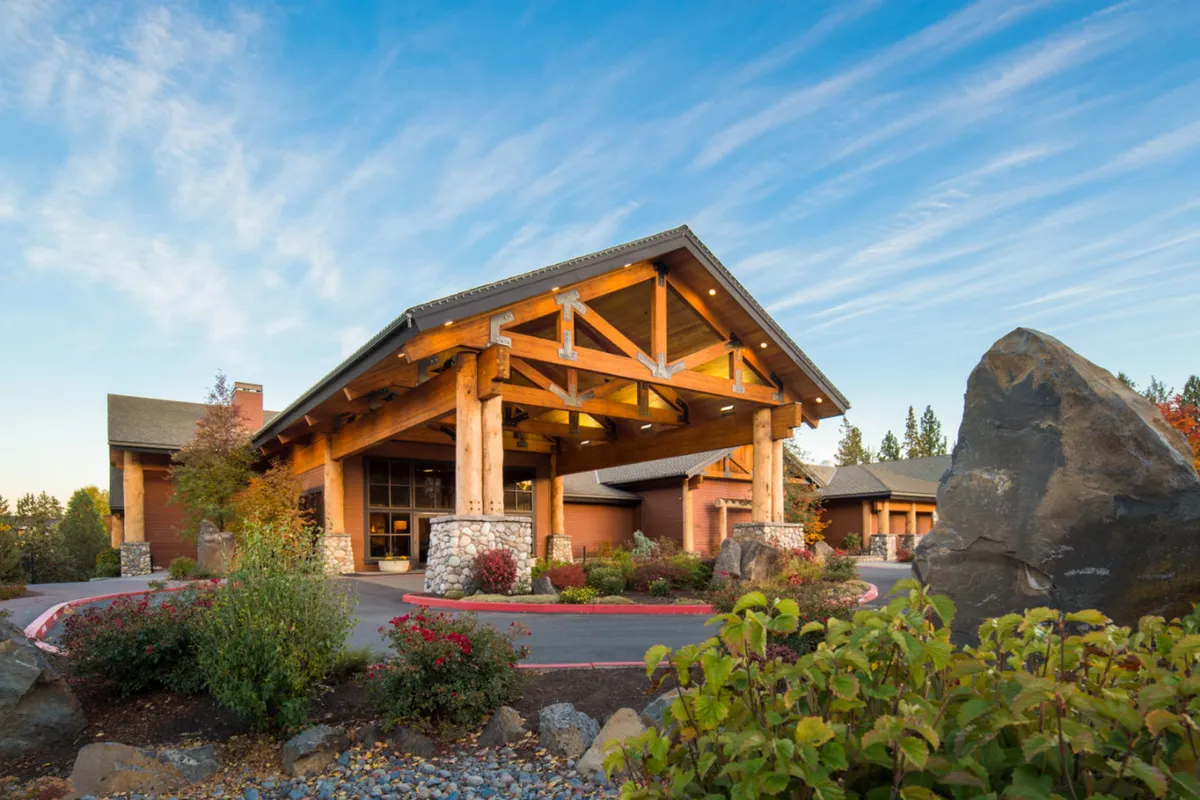An exterior view of Riverhouse Lodge’s dramatic wooden entrance and stonework, surrounded by landscaped gardens and set against a stunning sunset sky. The scene evokes gatherings, arrivals, or departures—moments when families connect and begin their adventures together. The mood is serene, emphasizing nature and relaxation.