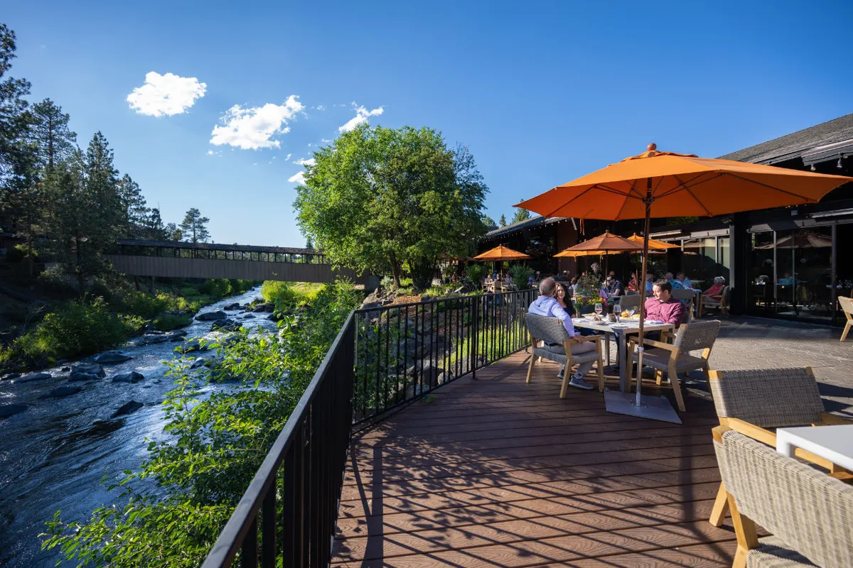 A lively scene set along the river featuring dining tables with orange umbrellas, guests socializing and enjoying meals outdoors, and lush green trees flanking the flowing water.