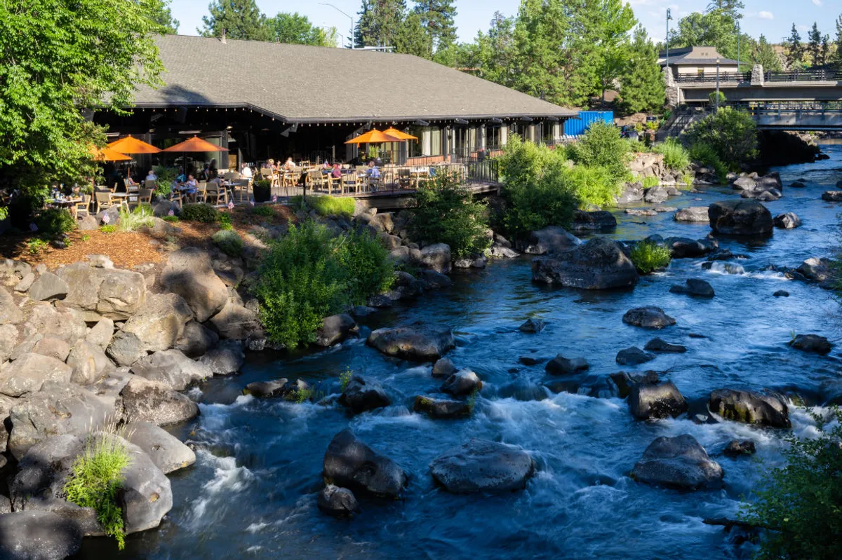 The image showcases a lively restaurant patio overlooking the flowing Deschutes River, framed by dramatic rock formations and lush greenery. Vibrant orange umbrellas shade tables where guests dine outdoors, relax by the water, and socialize as the sun sets. The inviting scene hints at live music events and the joy of seasonal food festivals, all in a serene, natural setting.