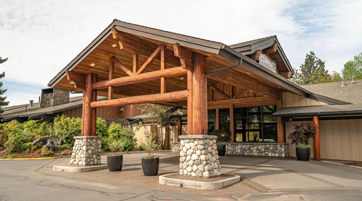 A welcoming view of the lodge's glass entrance framed by large wooden beams and stone pillars, surrounded by well-maintained greenery. The architectural details signal a modern, inviting space designed for arrivals and gatherings.