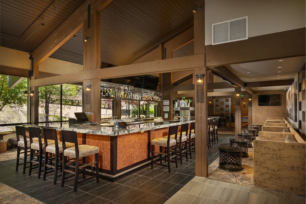 A stylish bar counter with high stools, a well-organized bottle display, and a relaxed seating area. The space is designed for comfort and interaction, with TVs positioned for watching games and atmosphere lighting that creates a welcoming mood. Guests can be seen enjoying drinks while engaging in conversation or sports viewing.
