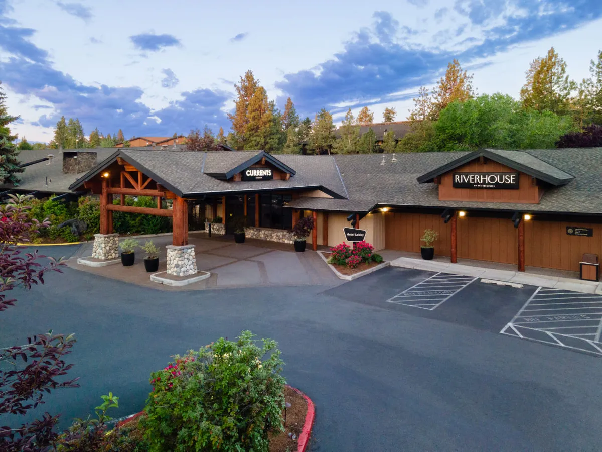 The image captures the Riverhouse Lodge entrance, framed by rustic wooden beams and stone accents. The signage for CURRENTS and RIVERHOUSE is visible, and lush landscaping surrounds the entryway. The scene is lively, with families and guests coming and going, set against a welcoming and relaxed mood. Sunset light adds warmth to the setting.